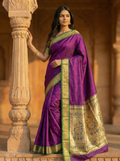 Woman in a purple saree with gold border standing against a stone wall.
