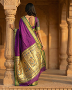 Woman in a traditional purple and gold saree standing in front of stone pillars.