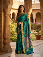 Woman in a teal saree with gold details standing in an outdoor setting with architectural columns and plants.