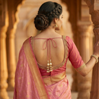 Woman in a traditional pink and gold saree with intricate designs, standing in a warm-toned architectural setting.