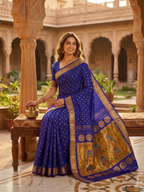 Woman in a blue saree with gold details sitting in an outdoor setting.