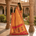Woman in a traditional yellow saree with red border standing in an outdoor setting with columns and greenery.